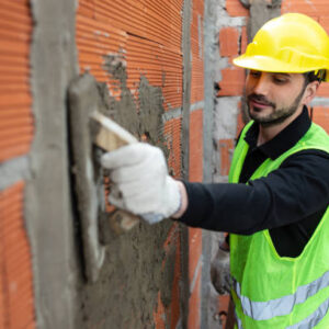 Male worker plastering cement on brick wall at construction site. A professional mason plastering a wall of a room. Construction worker wearing reflective clothing and hardhat.