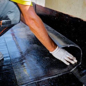 Worker in gloves prepare for melting, manually place and unroll rolled up, sheet of waterproofing installation over concrete. Residential building under construction placing water insulation